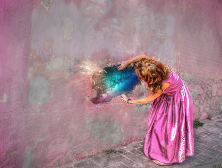 Pink coloured wall with woman in pink dress ripping open a hole to reveal another colour that looks like a galaxy